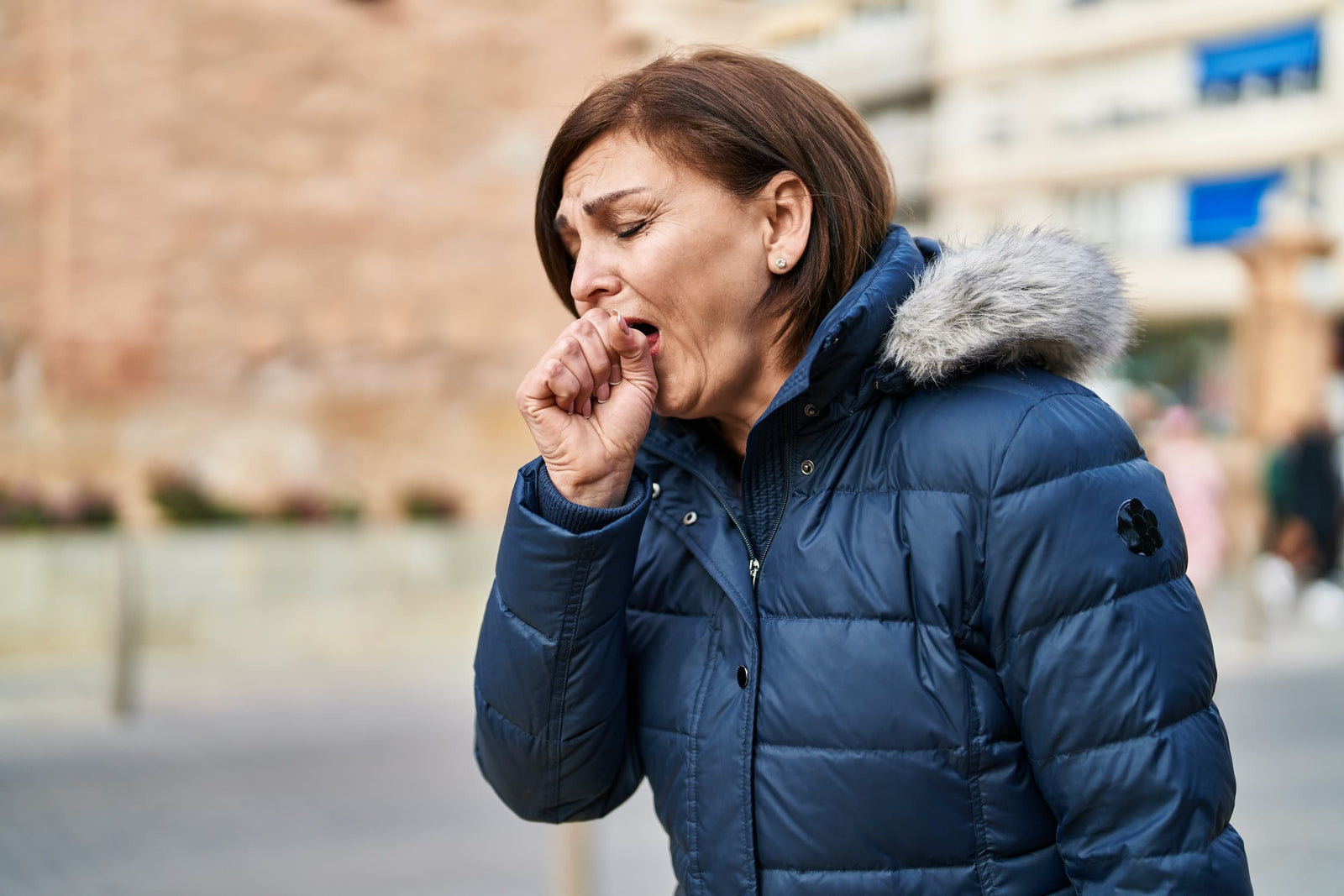 A woman in a thick, winter coat coughs while walking down the street.